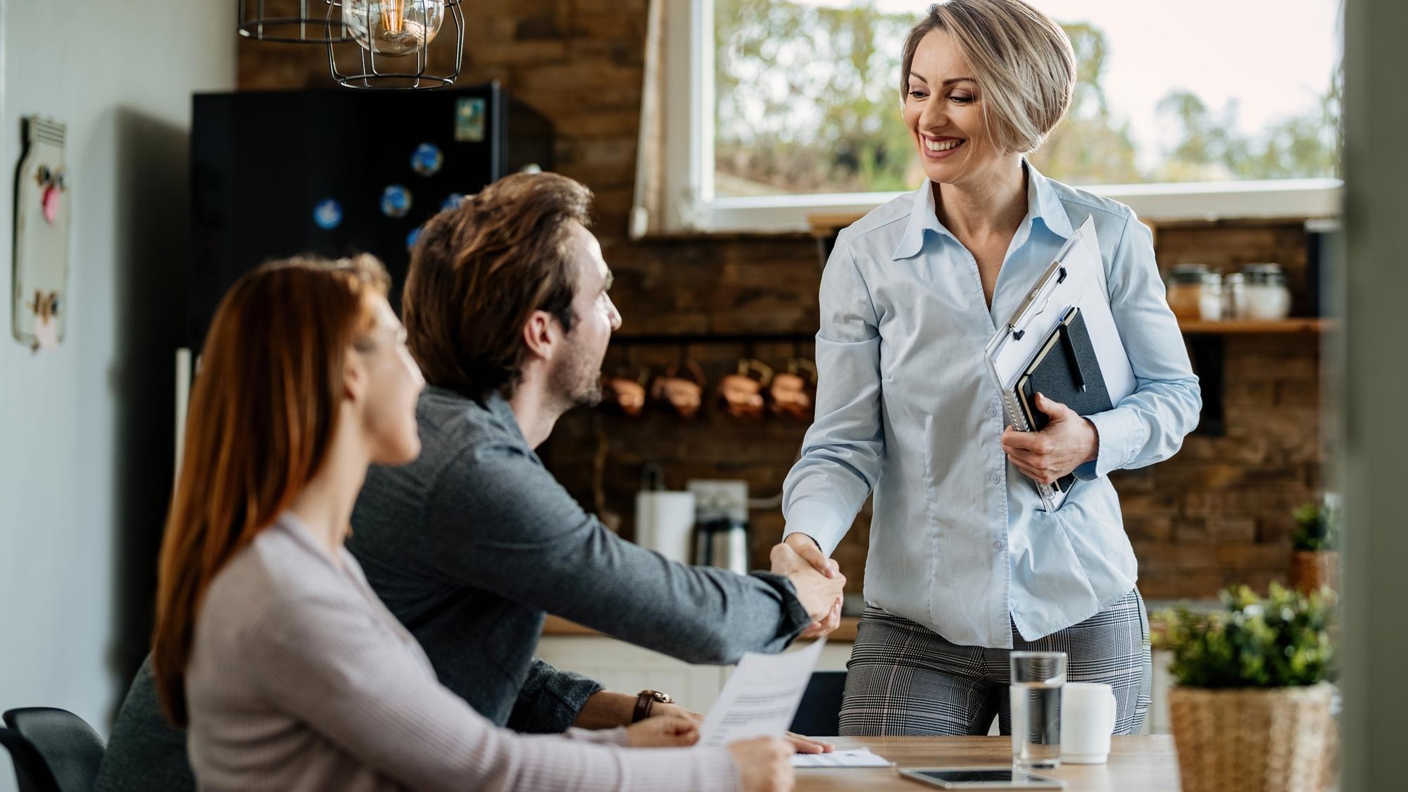 Happy insurance agent meeting with young couple and shaking hands with a man at their home