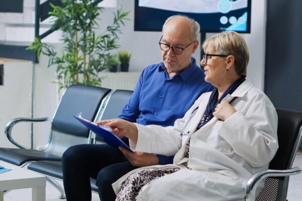 Two people in a waiting room, one of them is wearing a white coat and the other is wearing glasses.