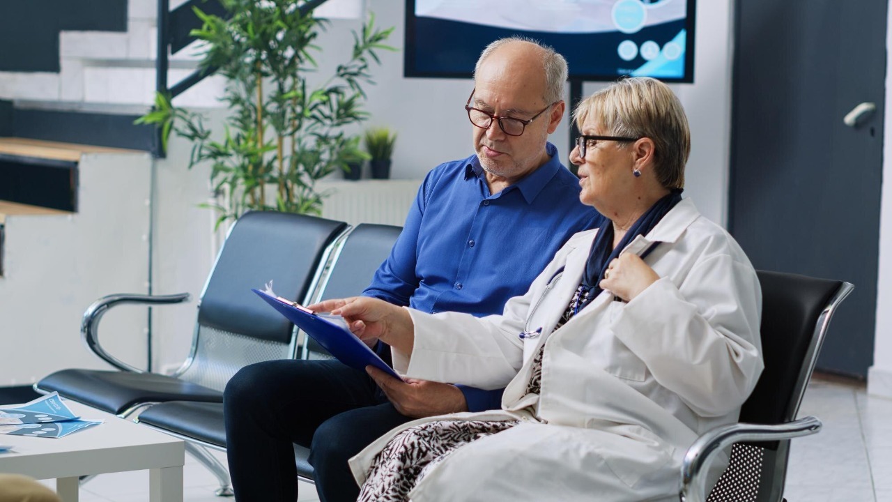 Two people in a waiting room, one of them is wearing a white coat and the other is wearing glasses.