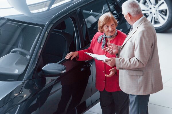 Old man in formal wear with notepad supporting woman in choosing automobile.