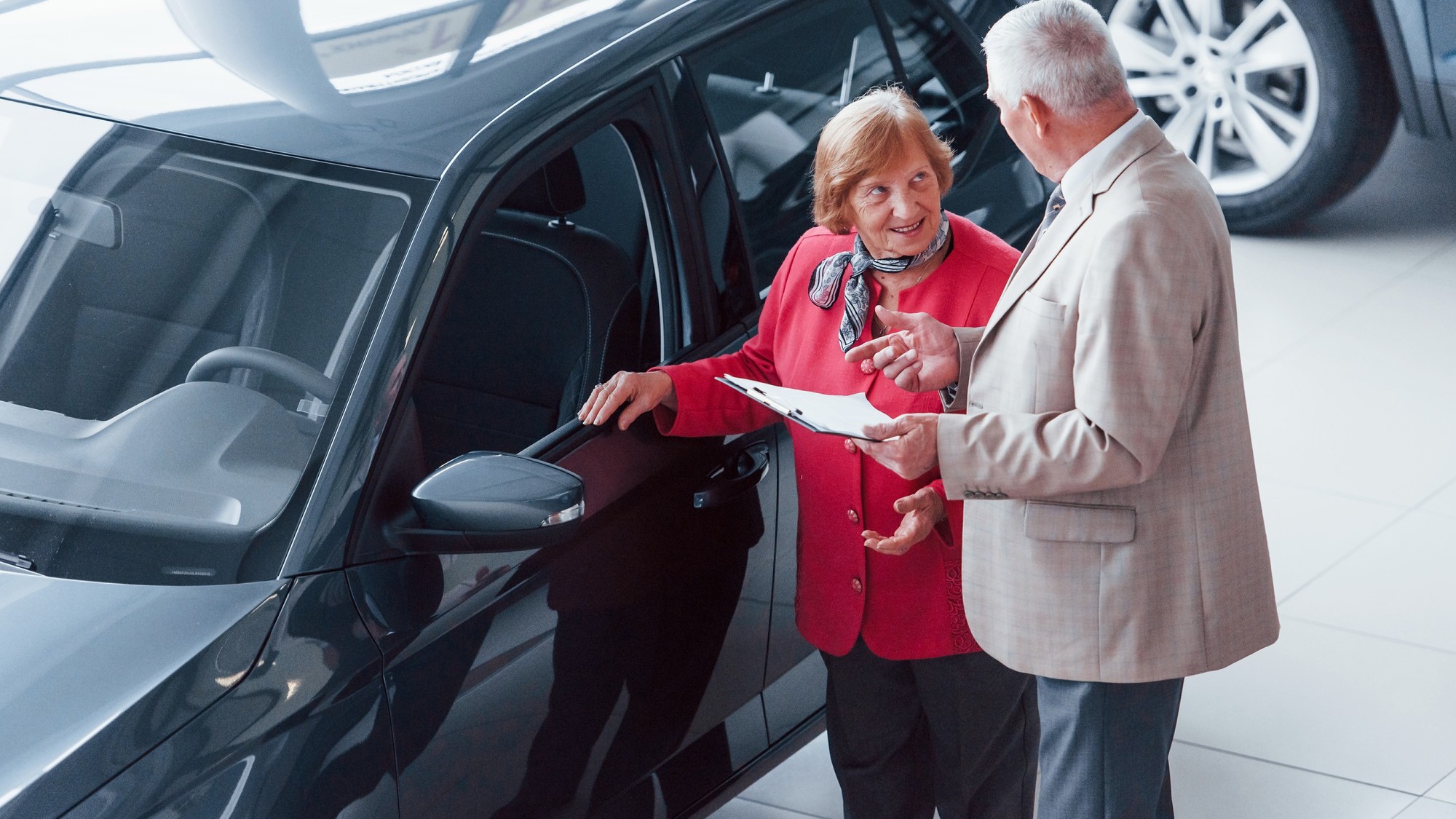 Old man in formal wear with notepad supporting woman in choosing automobile.