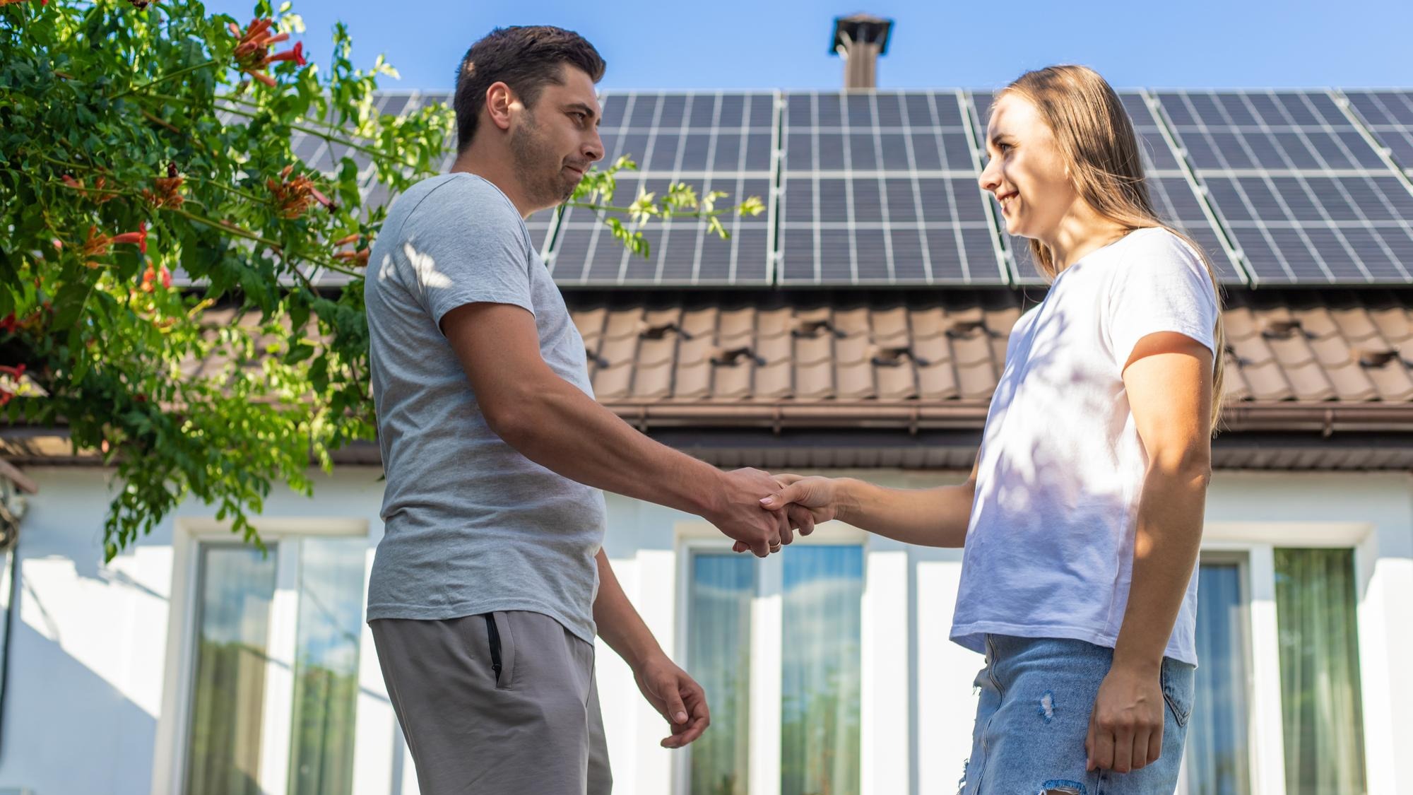 Smiling man and woman shaking hands in front of a house with solar panels Agreement