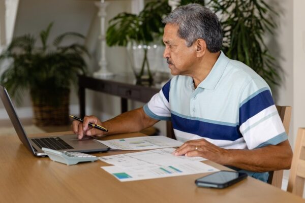 Senior biracial man doing paperwork and using laptop in dining room. Finance, retirement, domestic life, communication and senior lifestyle, unaltered.