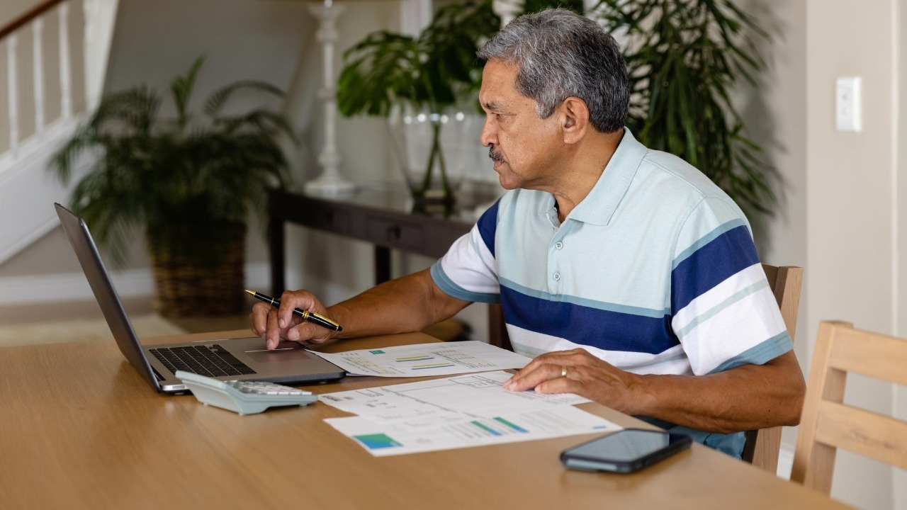 Senior biracial man doing paperwork and using laptop in dining room. Finance, retirement, domestic life, communication and senior lifestyle, unaltered.