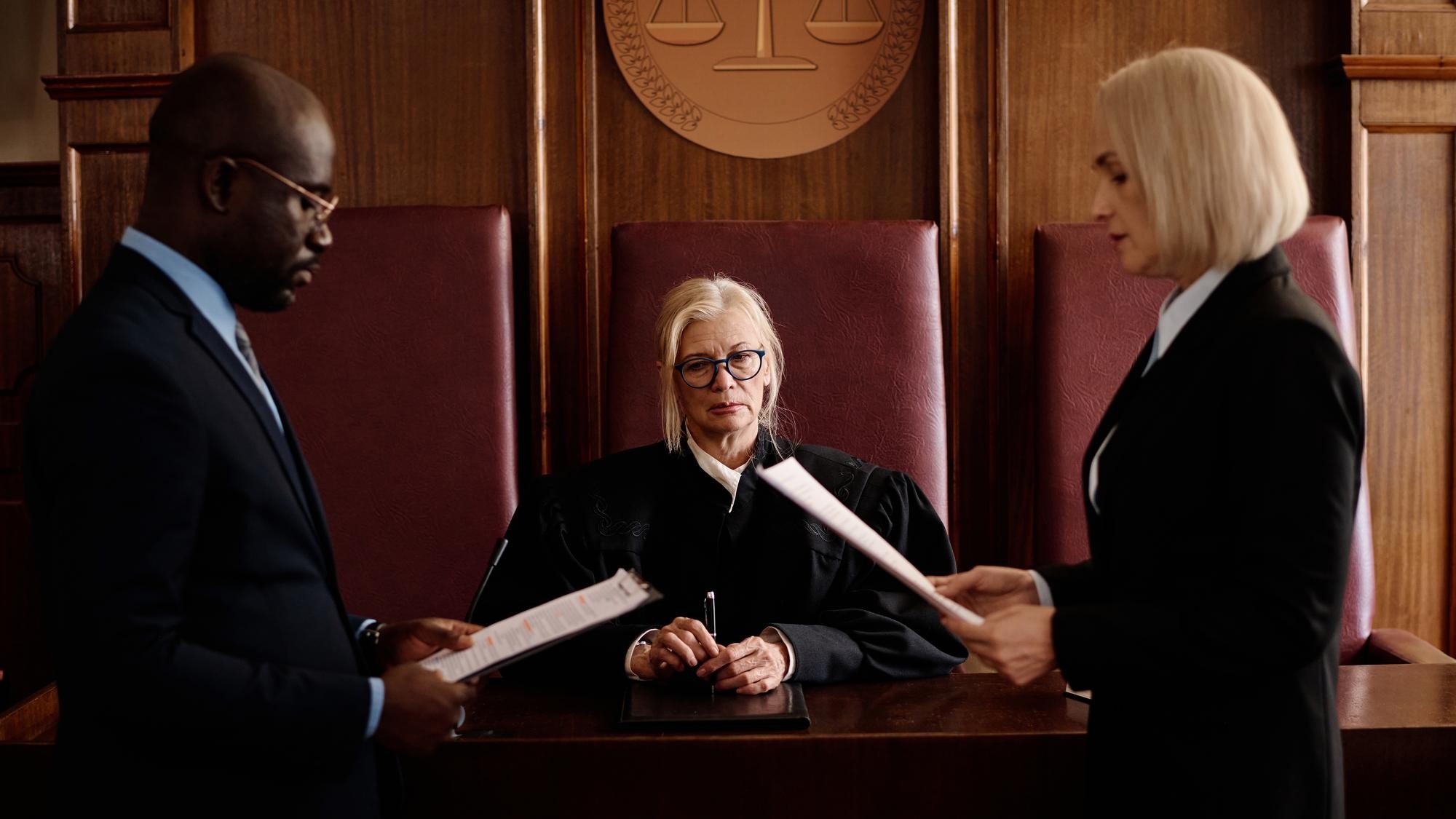 Confident mature female judge sitting by desk between prosecution and defense sides