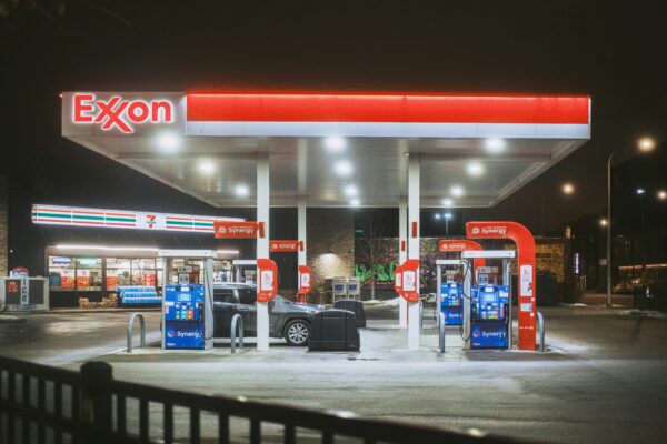 A brightly lit Exxon gas station and 7-Eleven store at night, featuring fuel pumps and a parked car.