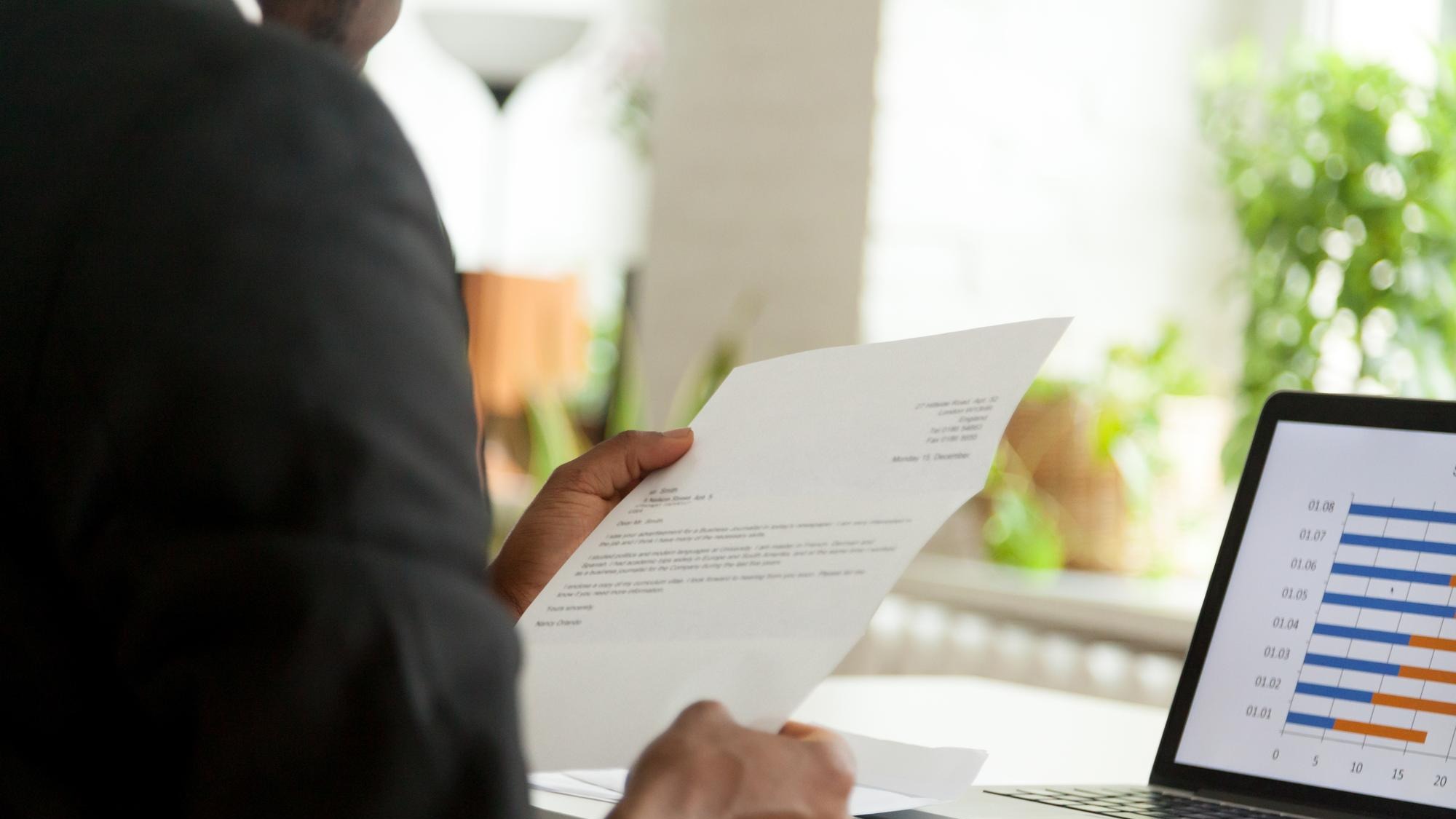 Africanamerican hr manager or employer reading cover letter from job vacancy applicant black businessman holding business mail checking paper correspondence at workplace over the shoulder view