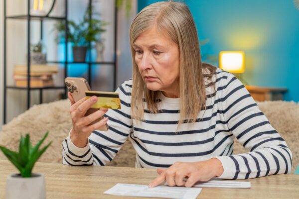 Mature woman using credit card and smartphone to transfer money pay bills sitting on table at home