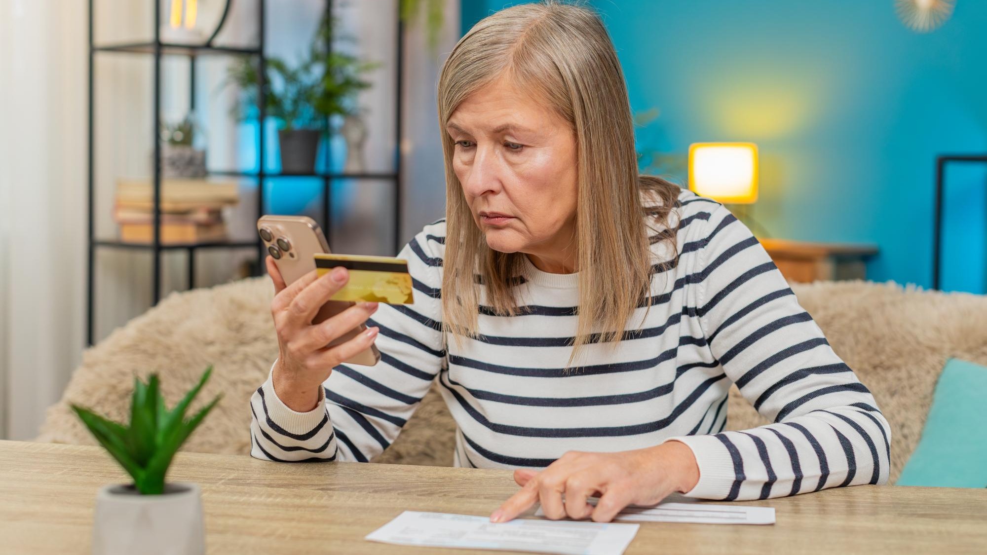 Mature woman using credit card and smartphone to transfer money pay bills sitting on table at home