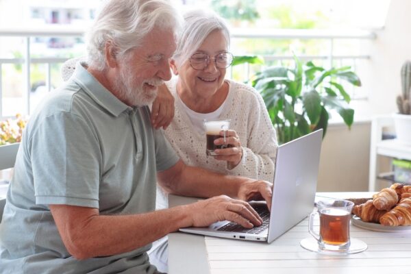 Smiling senior retired couple browsing with laptop while enjoy breakfast together sitting outdoors