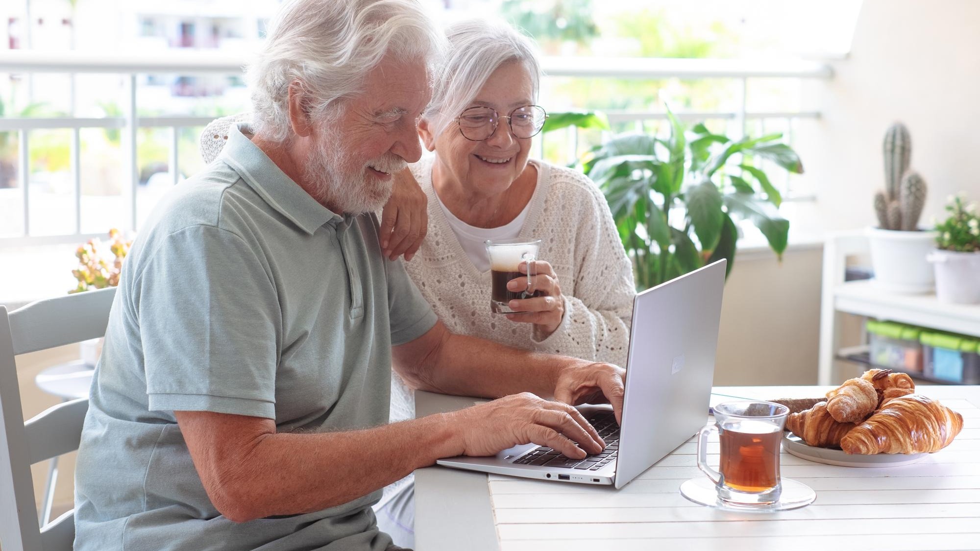 Smiling senior retired couple browsing with laptop while enjoy breakfast together sitting outdoors