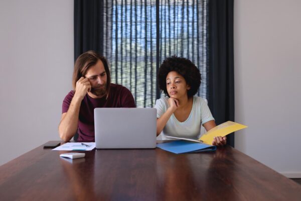 Diverse couple sitting at table working from home, using laptop and checking documents