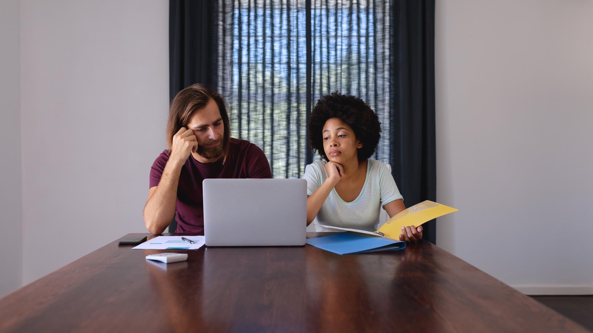Diverse couple sitting at table working from home, using laptop and checking documents
