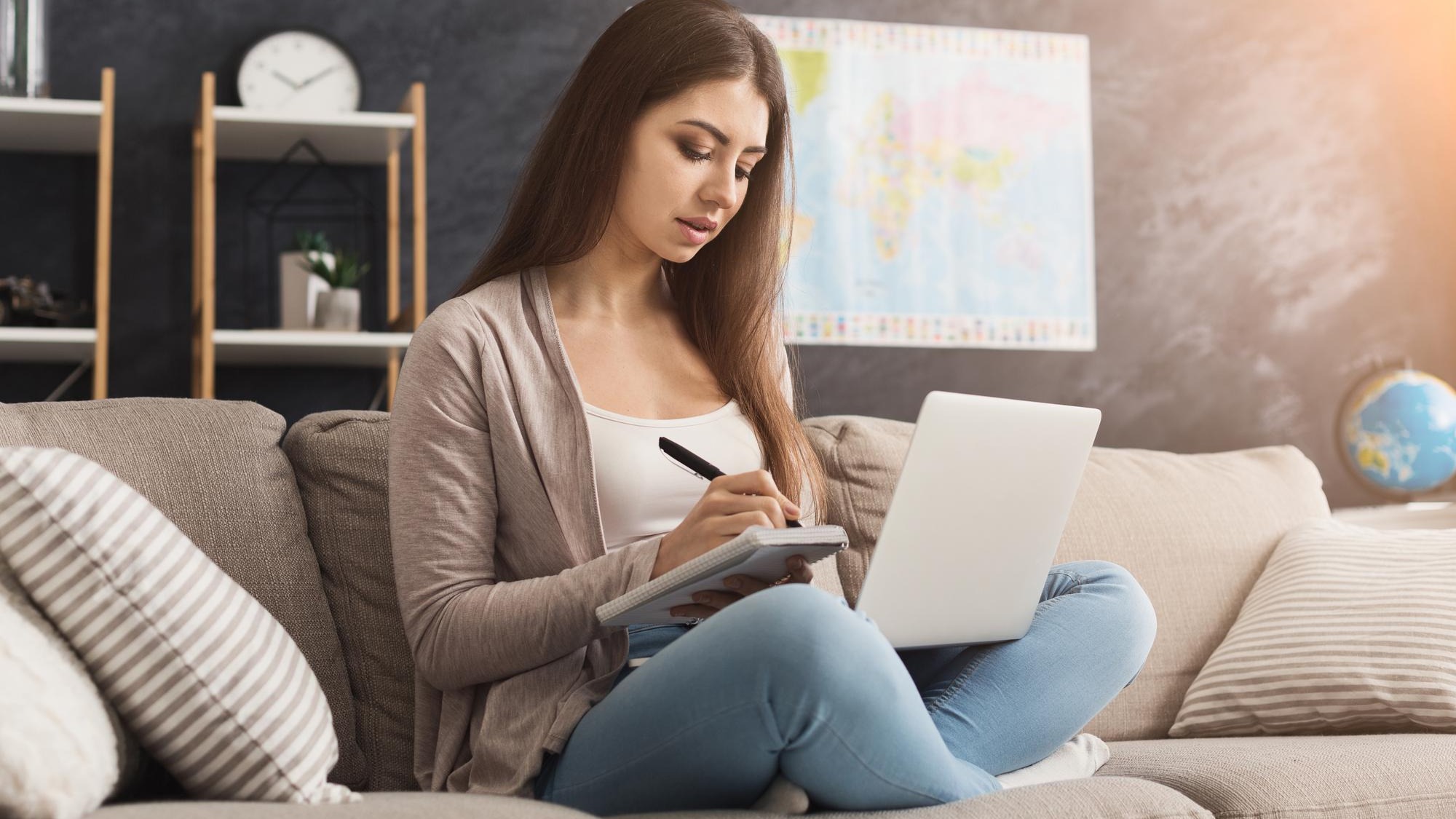 Thoughtful woman using laptop and writing notes to notebook at home while sitting on couch, copy space