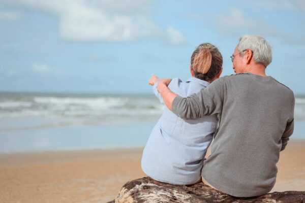A retired couple sitting at the sea, a woman pointing a finger for a man to watch something on the rocks and holding hands at the beach in the morning, life insurance plans at retirement concepts.