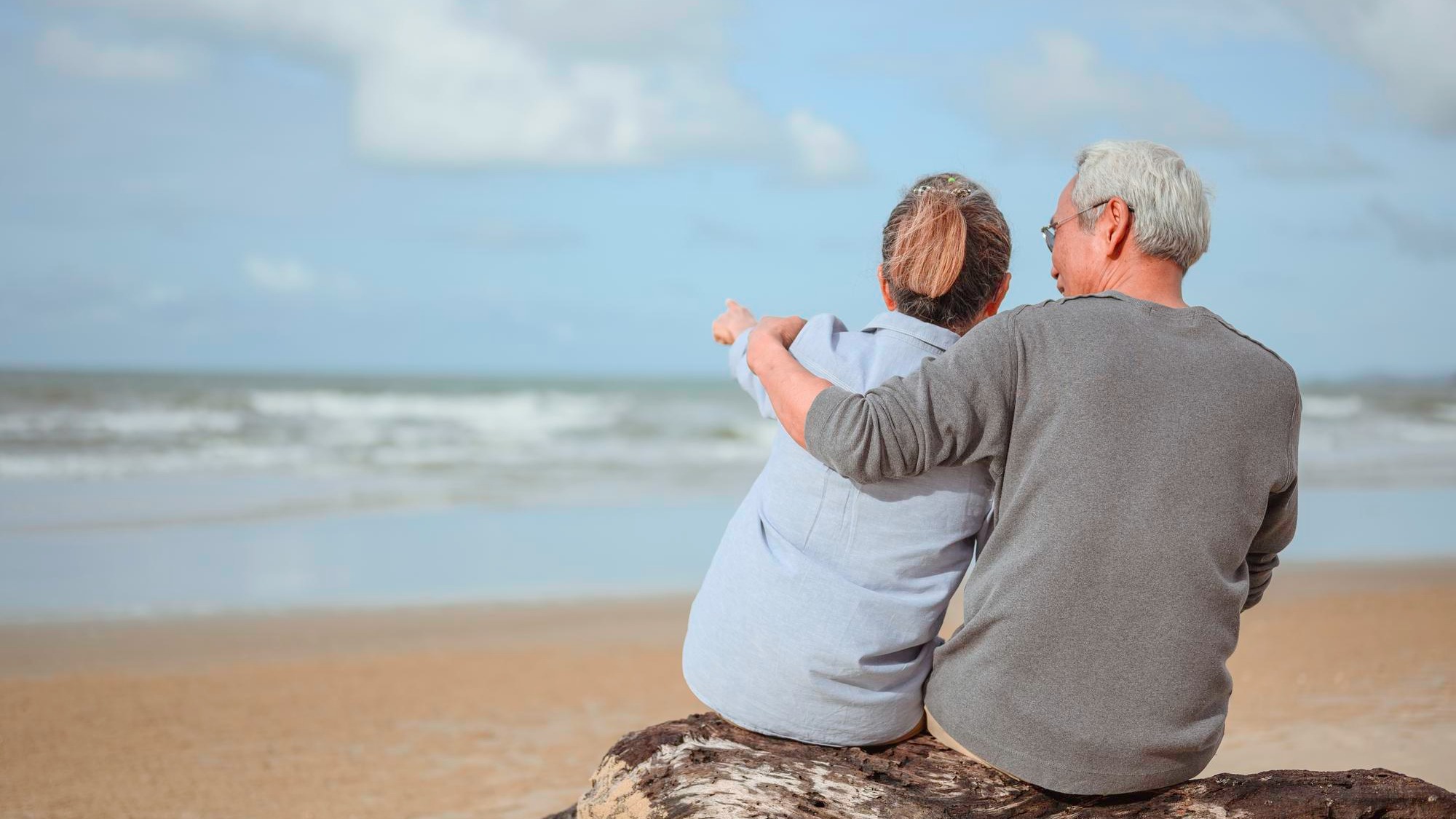 A retired couple sitting at the sea, a woman pointing a finger for a man to watch something on the rocks and holding hands at the beach in the morning, life insurance plans at retirement concepts.