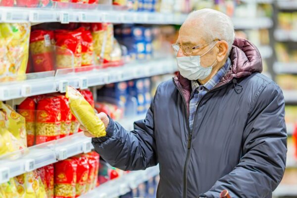Old senior european man wearing protective facial mask looking at groceries in the supermarket. Shopping during COVID-19 concept.