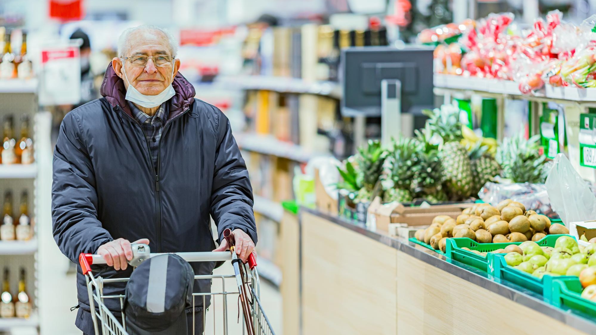 Old senior european man wearing protective facial mask pushing shopping cart in the supermarket. Shopping during COVID-19 concept.