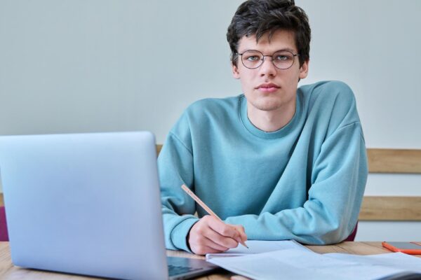 Young guy college student sitting at desk in classroom using laptop looking at camera