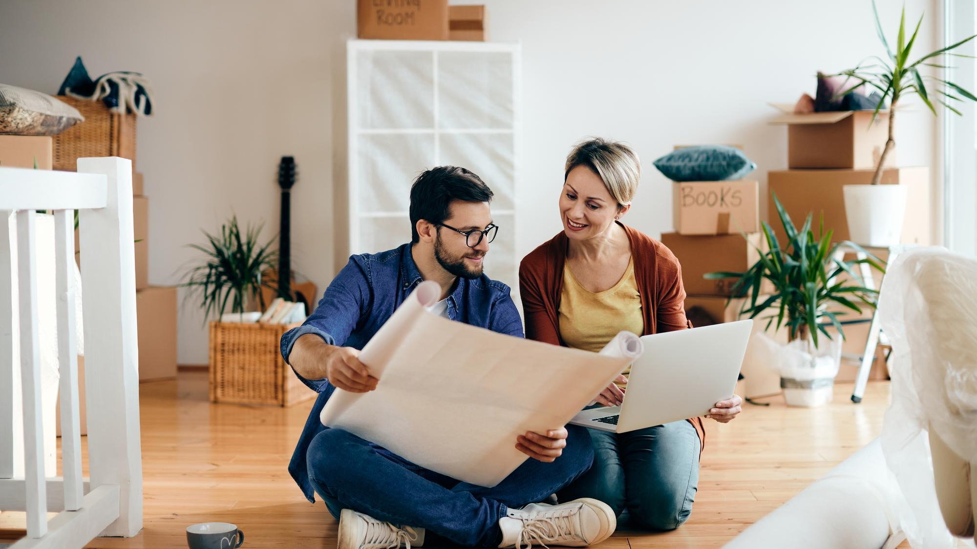Happy couple examining blueprints and using laptop at their new apartment