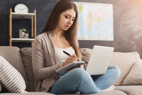 Thoughtful woman using laptop and writing notes to notebook at home while sitting on couch, copy space