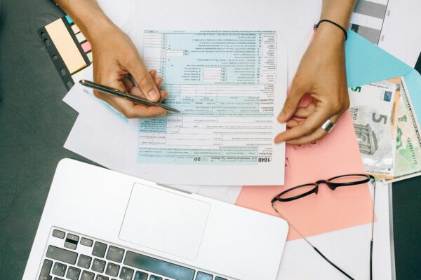 Hands writing on tax documents with laptop, glasses, and currency on desk.
