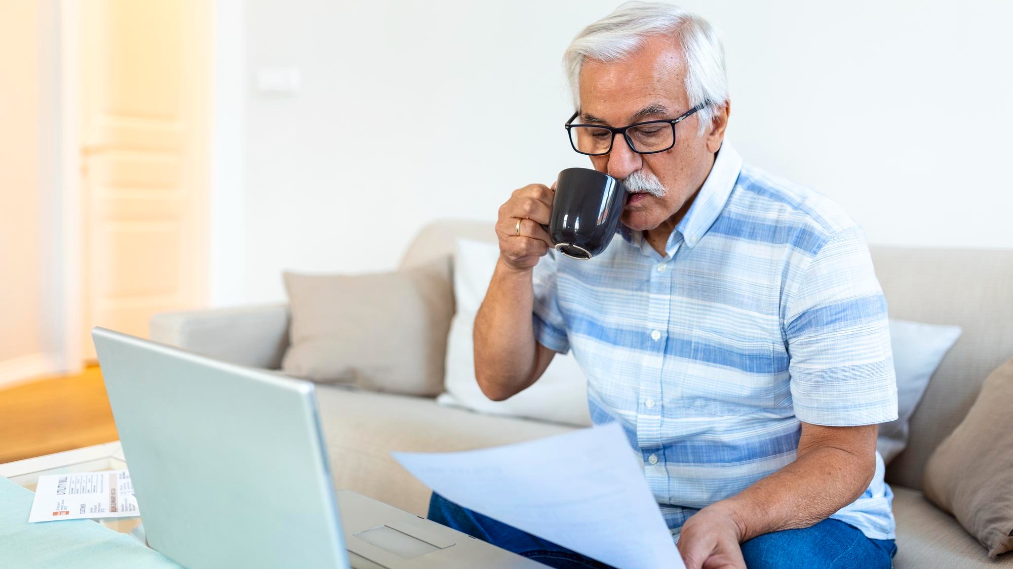 Focused greyhaired elderly man sit on couch reading bank notifications calculating domestic expenditures concentrated modern mature male consider financial paperwork pay bills on laptop online