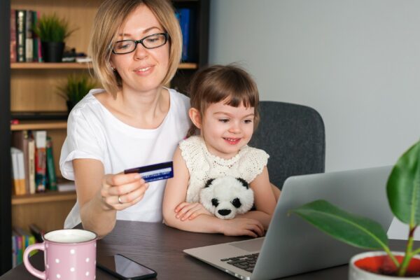 Mother holding a credit card with her little daughter sitting near looking at the laptop. Shopping online concept.