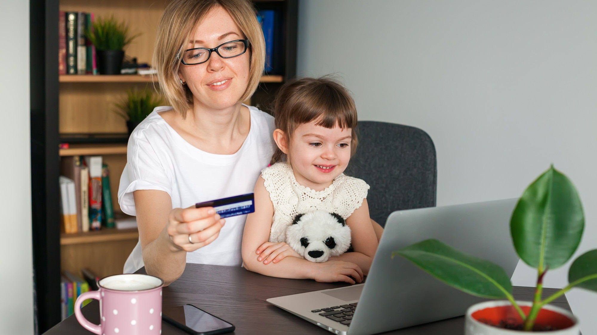 Mother holding a credit card with her little daughter sitting near looking at the laptop. Shopping online concept.