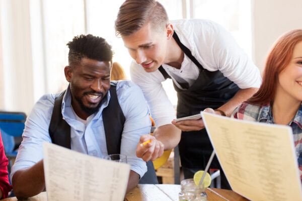 service and people concept - group of happy international friends with menu and drinks and waiter with notepad receiving order at bar or restaurant