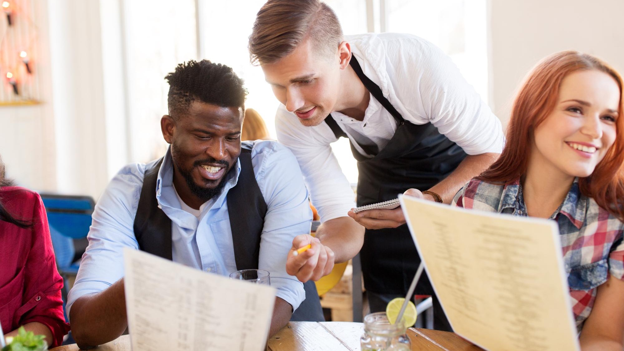 service and people concept - group of happy international friends with menu and drinks and waiter with notepad receiving order at bar or restaurant