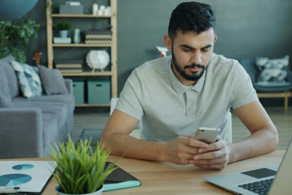 Man using smartphone at desk with laptop and charts.