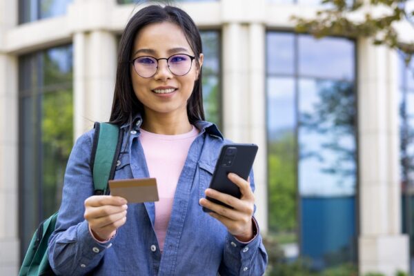 An asian woman stands outside holding a credit card and smartphone with a smile