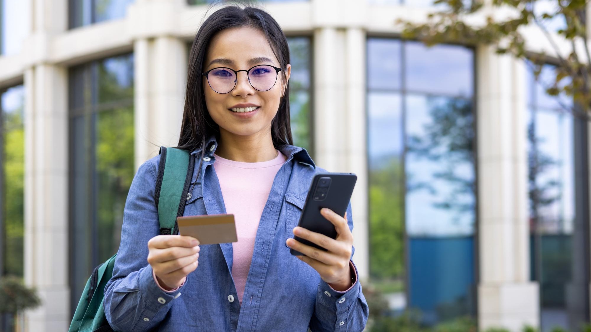 An asian woman stands outside holding a credit card and smartphone with a smile
