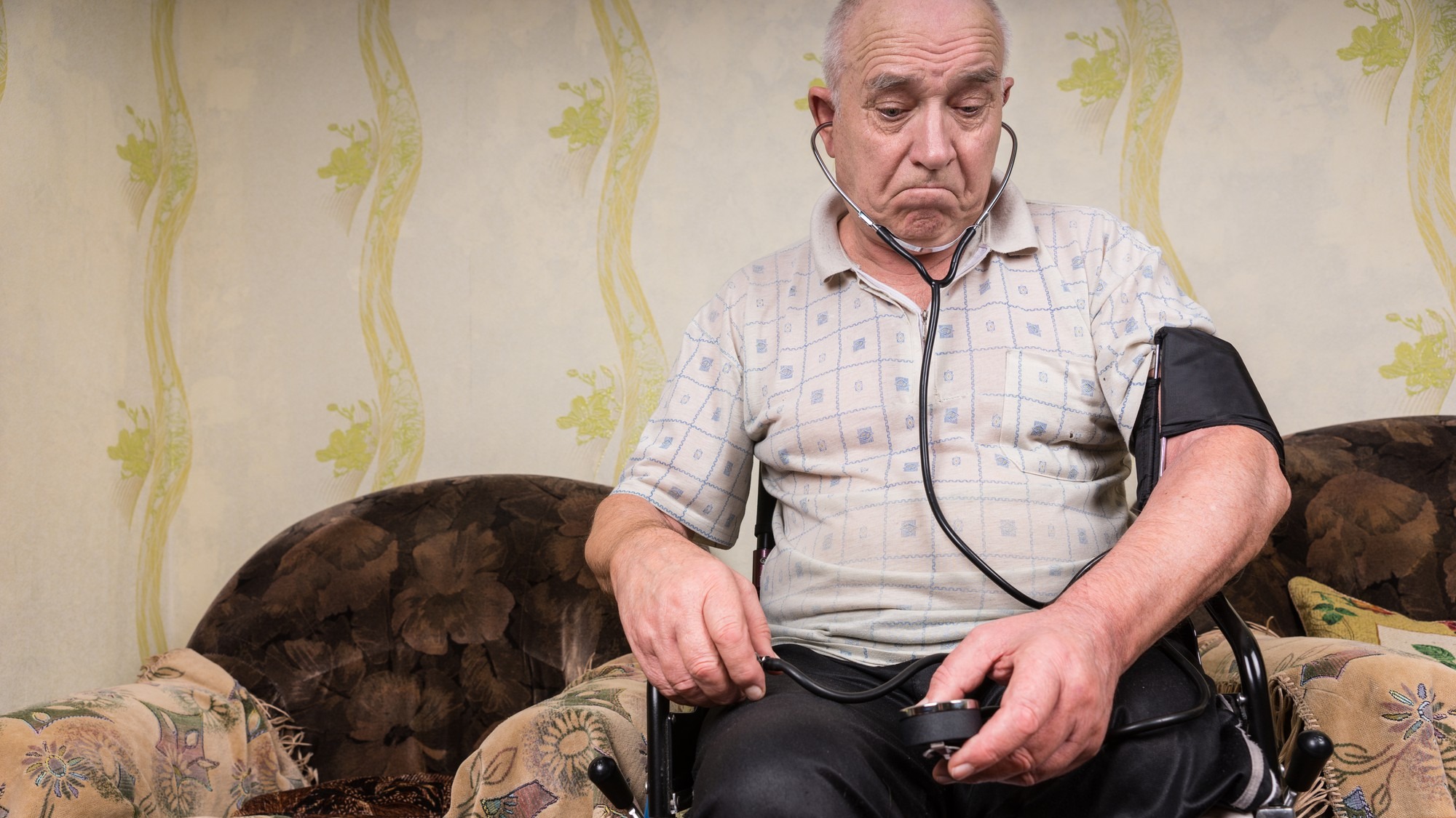 Thoughtful Old Bald Man with Blood Pressure Apparatus, Sitting on his Wheelchair In the Living Room with his Medicines on top of the Table.