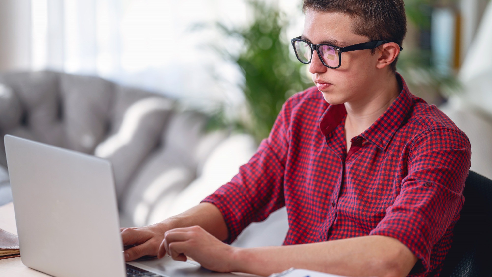 Brooding teen guy in glasses, listens to lecture, does in notebook, using laptop