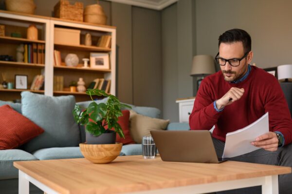 Focused businessman with documents reading information online over laptop on table at home office