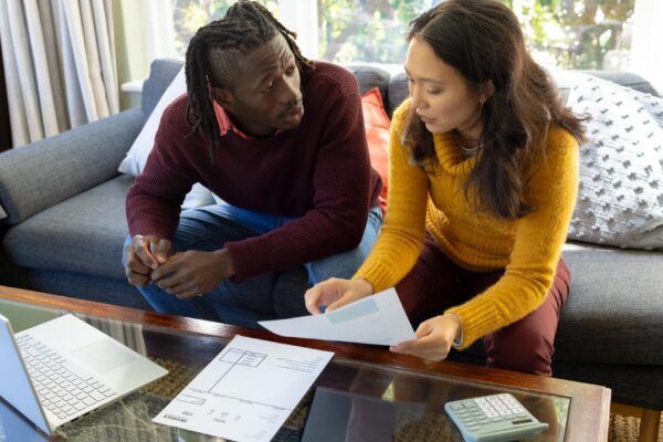 Serious diverse couple using laptop sitting on couch discussing bills and finances in living room