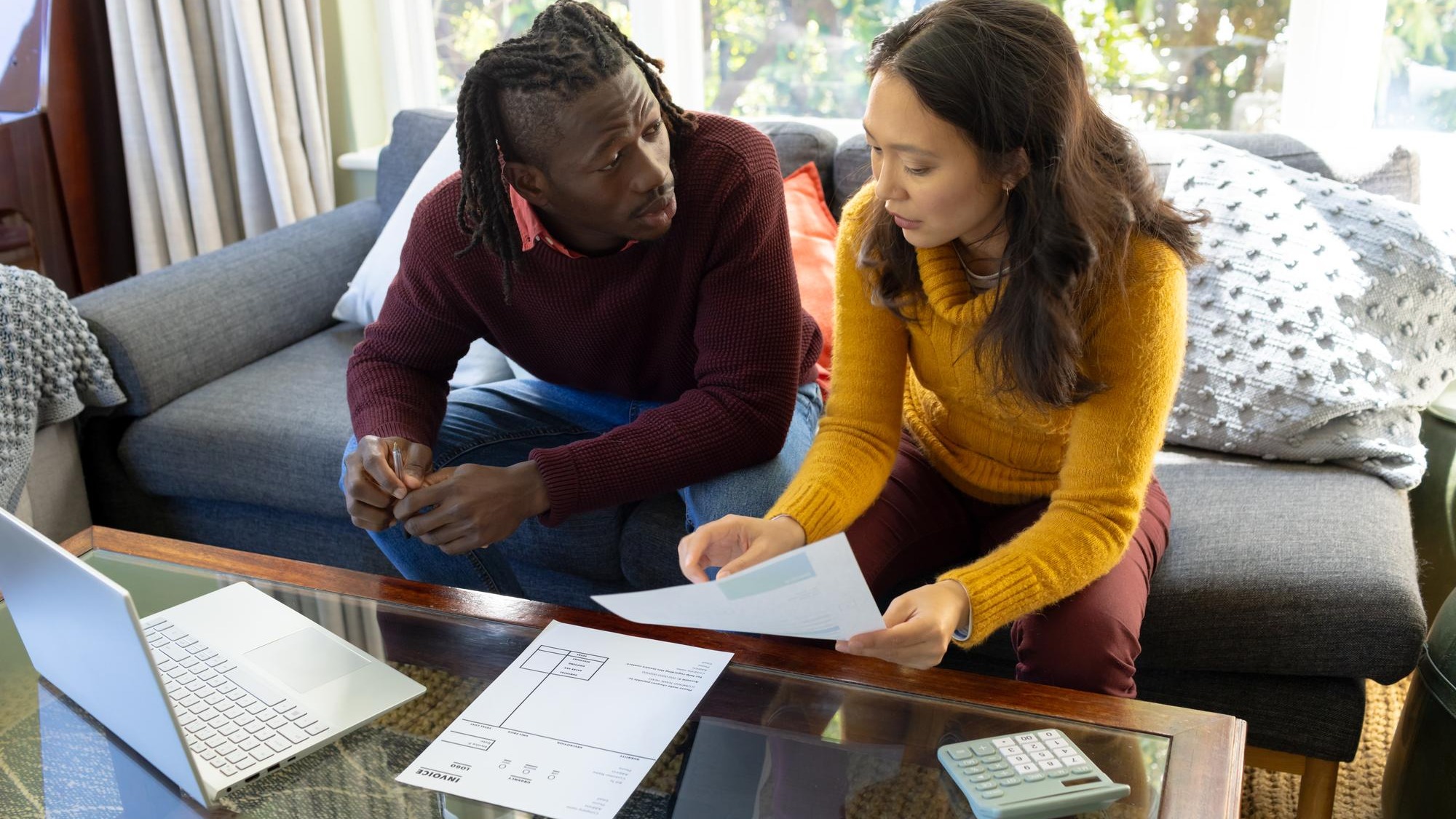 Serious diverse couple using laptop sitting on couch discussing bills and finances in living room