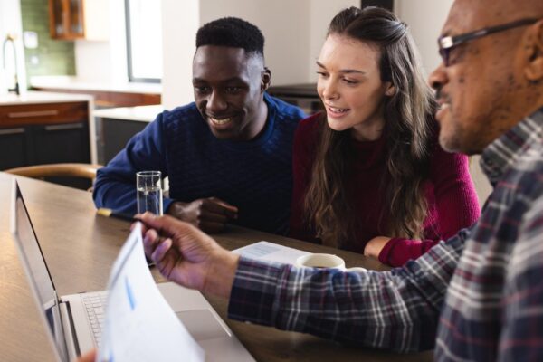 Diverse couple and biracial male financial advisor using laptop and discussing documents at home