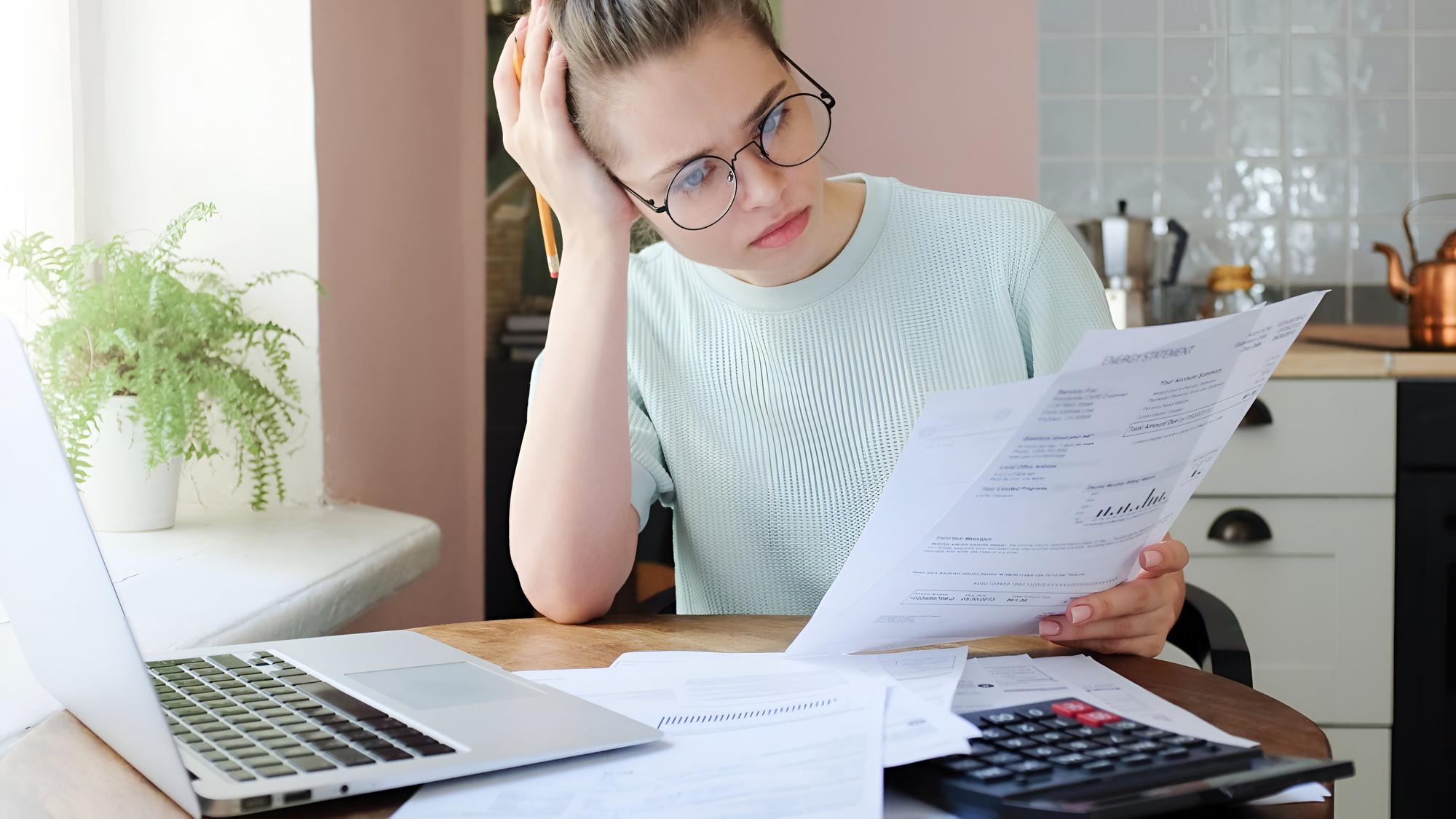 Young frustrated unhappy tired woman with financial troubles sitting at kitchen table with papers calculator and laptop computer