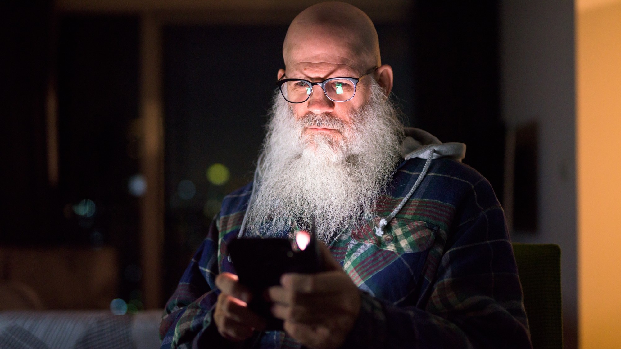 Mature bald bearded hipster man using phone at home in the dark