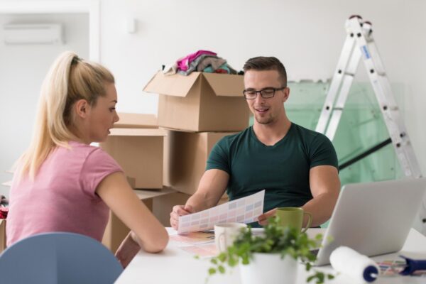 Young couple moving in a new home. Man and woman at the table using notebook laptop computer and plans with boxes around them