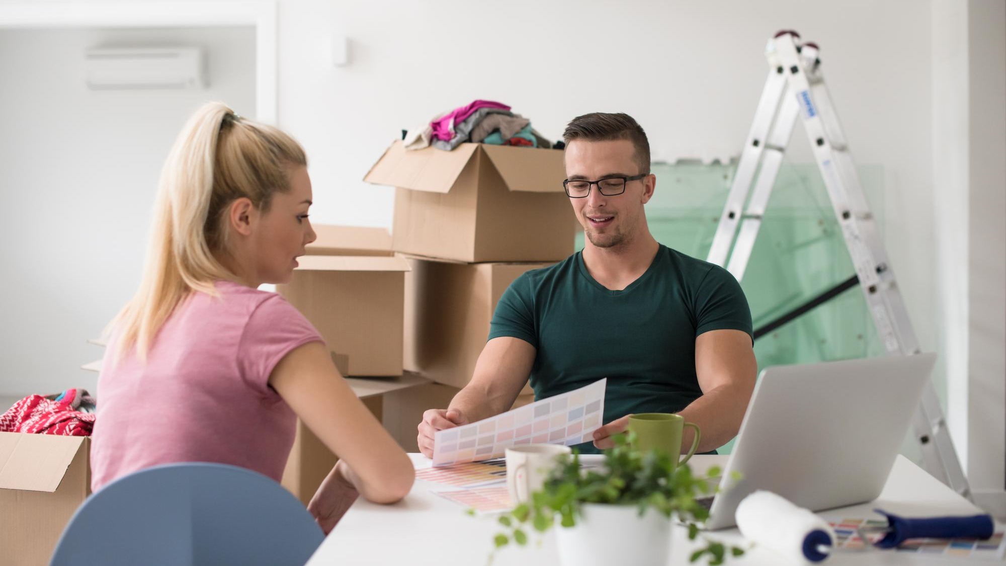 Young couple moving in a new home. Man and woman at the table using notebook laptop computer and plans with boxes around them