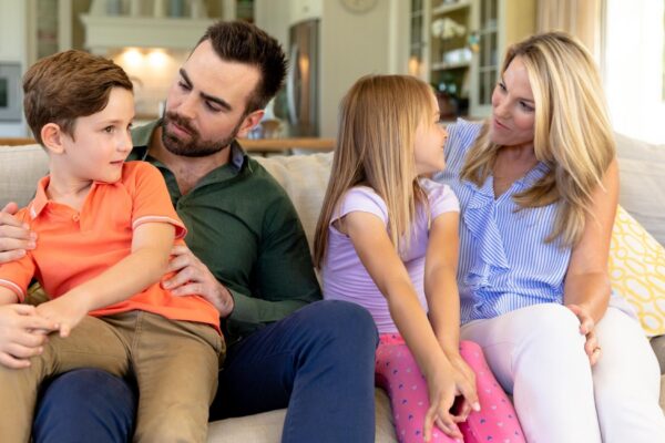 A Caucasian couple at home in the living room sitting on the couch, the father talking to their son, sitting on his knee, and the mother tallking to their daughter, sitting between her parents