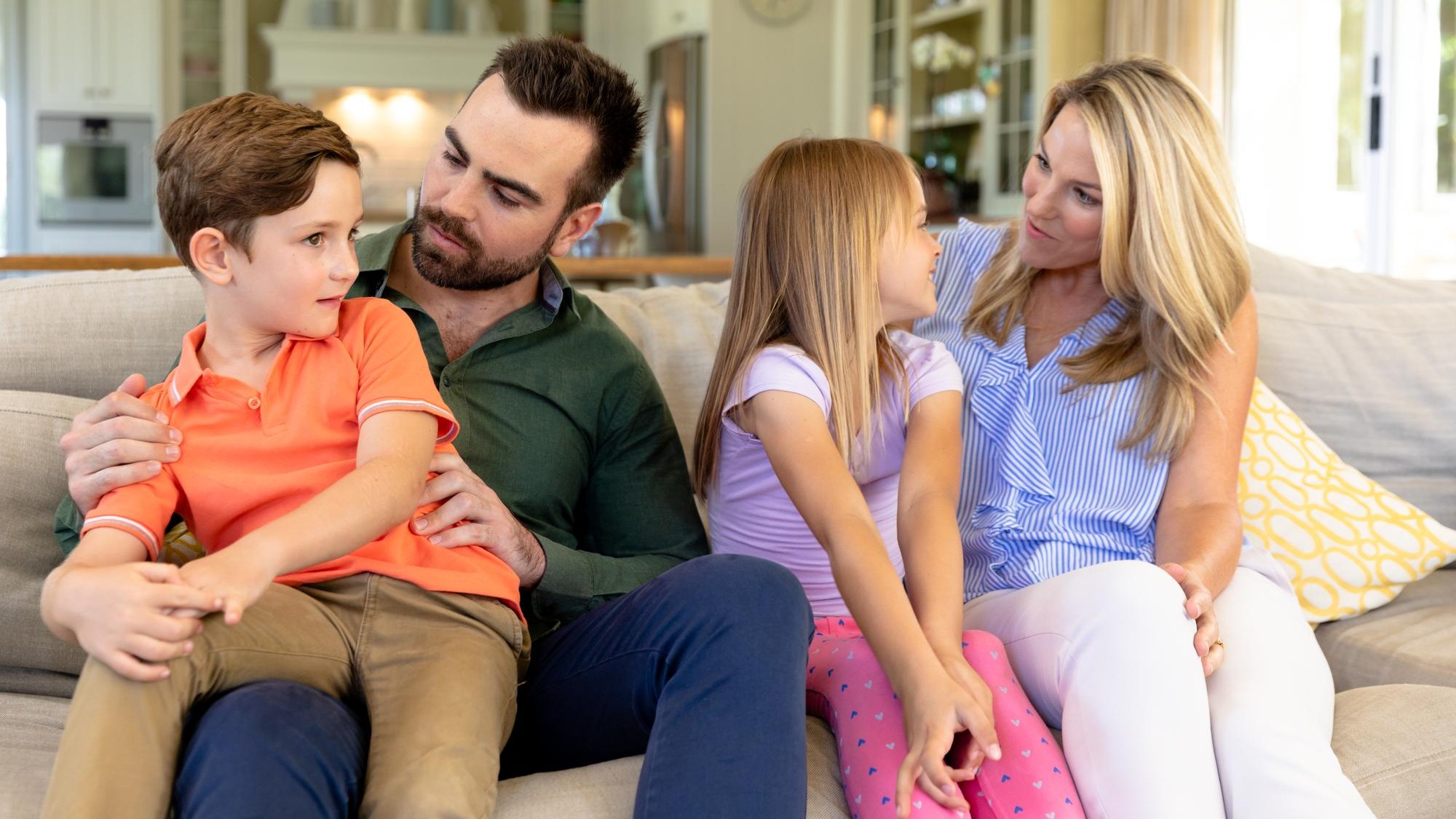 A Caucasian couple at home in the living room sitting on the couch, the father talking to their son, sitting on his knee, and the mother tallking to their daughter, sitting between her parents