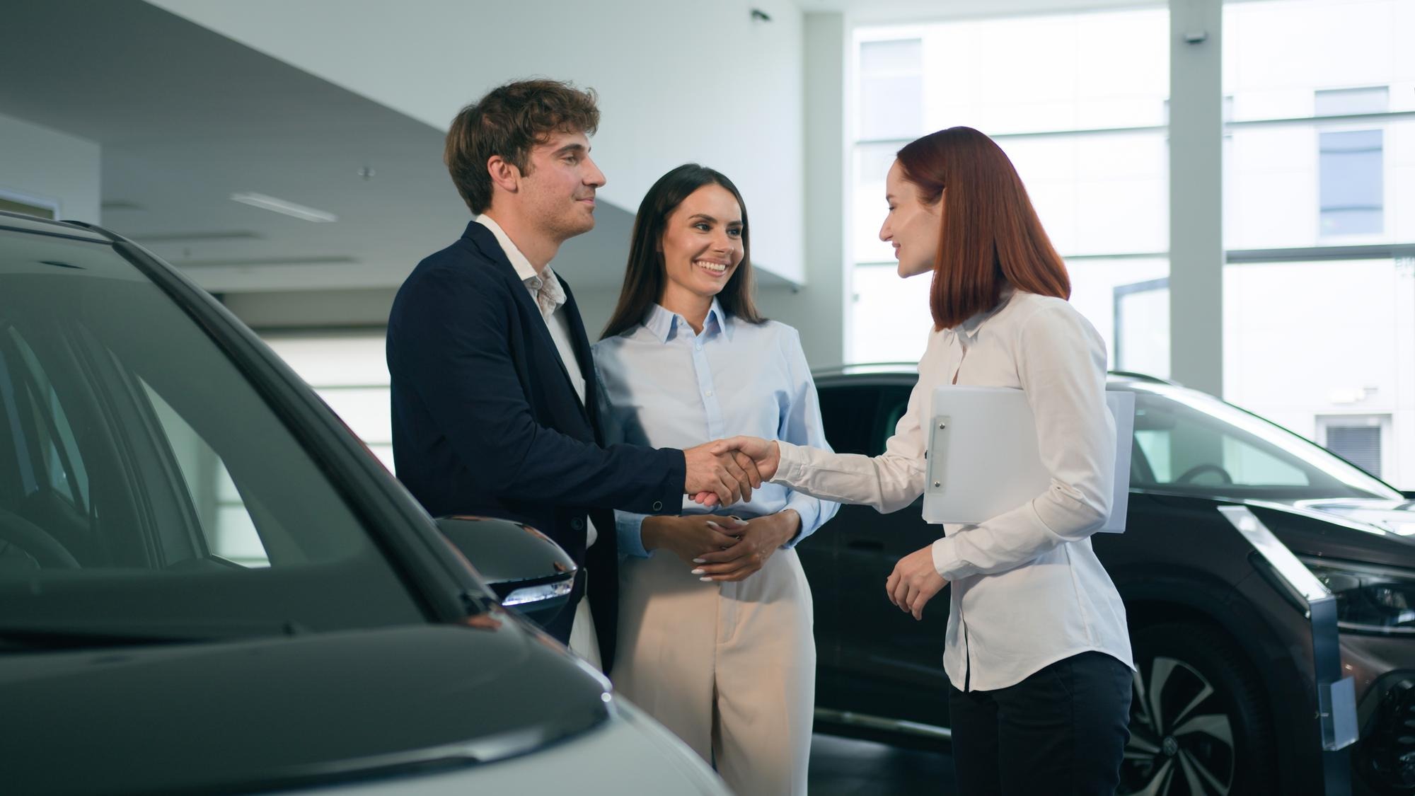 Caucasian couple clients buyers conclude agreement deal buy buying new car auto automobile in showroom rental service rent transport shaking hands with saleswoman woman dealer seller trader handshake