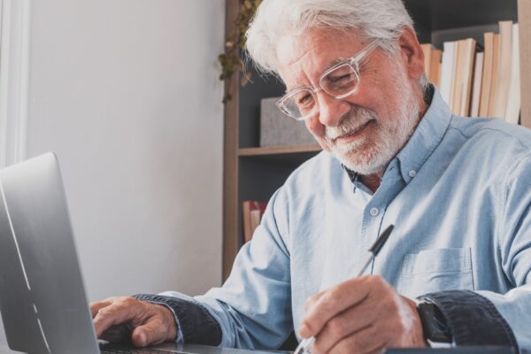 Happy old caucasian businessman smiling working online watching webinar podcast on laptop and learning education course conference calling make notes sit at work desk, elearning concept