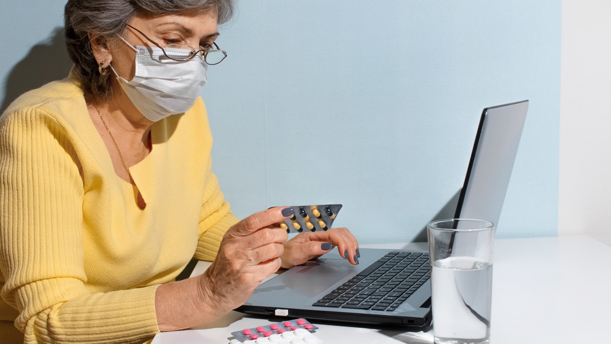 Senior woman with glasses and medical mask reading instructions of medicine. Elderly woman using laptop at home. Concept for online consultation with doctor, new normal.