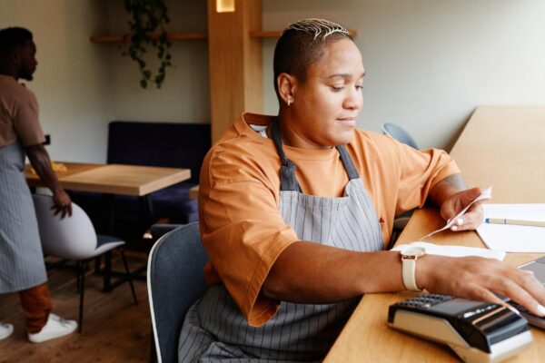 Young african american woman wearing apron sitting at table in front of laptop in modern cafe taking...